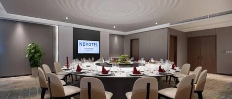 A conference room with a round table, beige chairs, and a projector screen displaying 'NOVOTEL'.