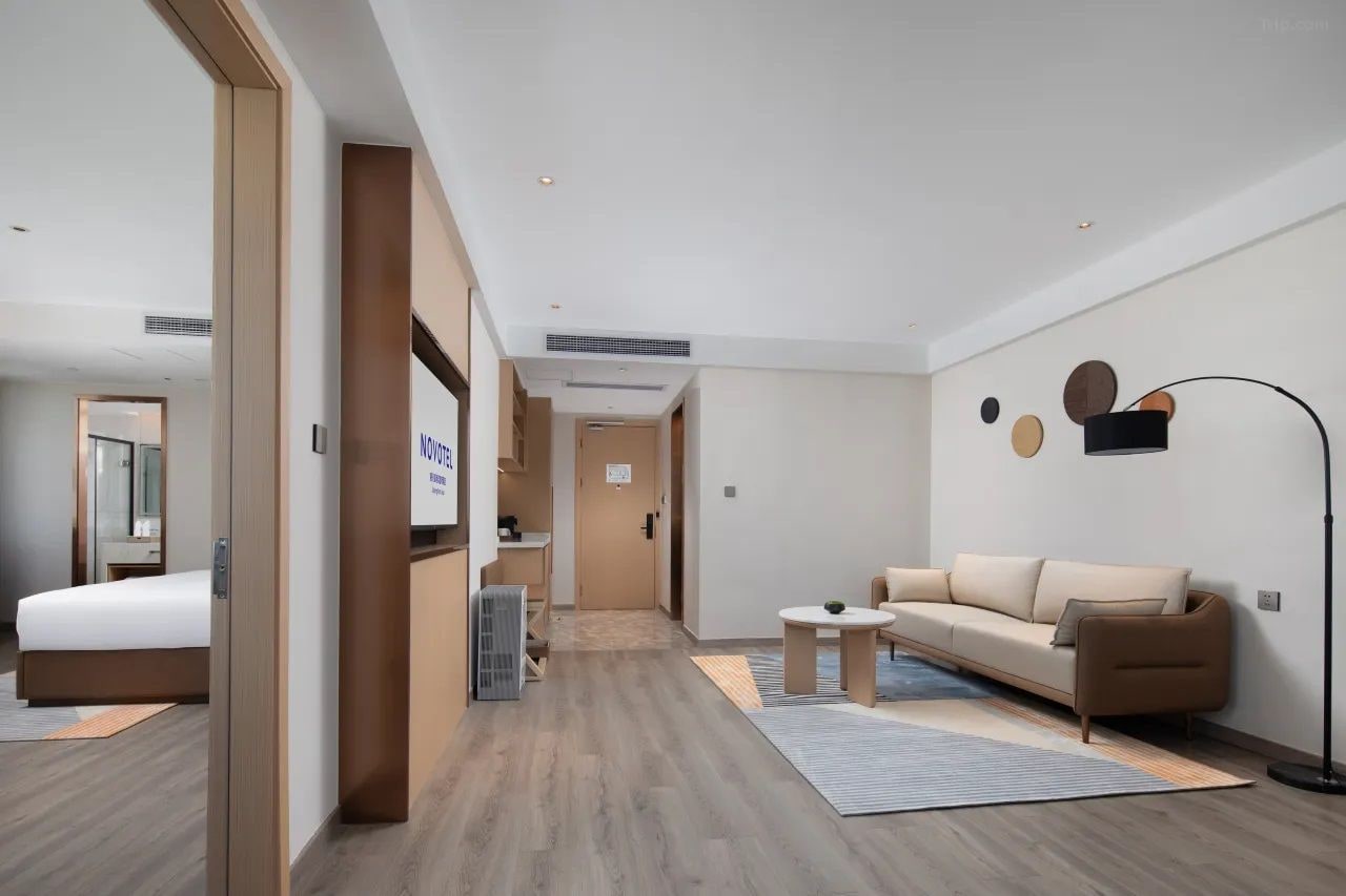 Una habitación de hotel moderna con una cama, un sofá, una mesita y una lámpara de pie. Se ve un pasillo que conduce a otra habitación.