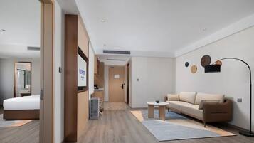 Una habitación de hotel moderna con una cama, un sofá, una mesita y una lámpara de pie. Se ve un pasillo que conduce a otra habitación.