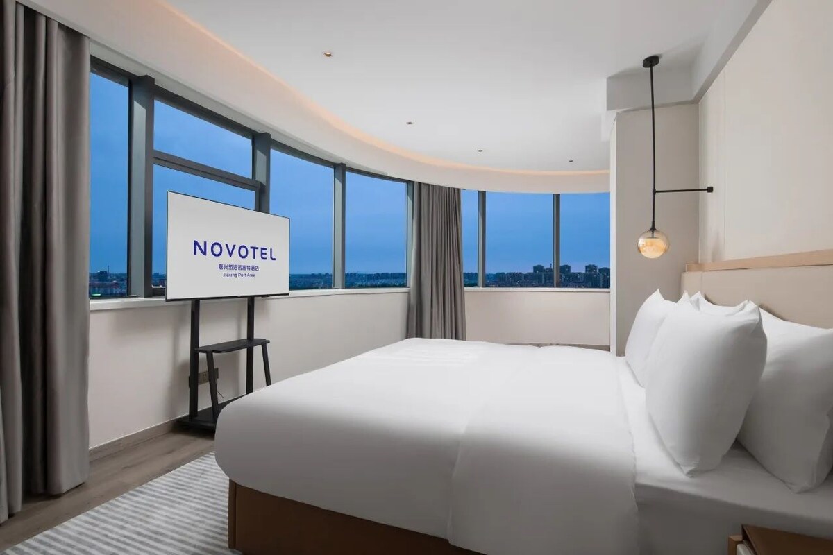 Una habitación de hotel moderna con una cama grande, un televisor sobre un mueble y vistas a la ciudad a través de amplios ventanales.