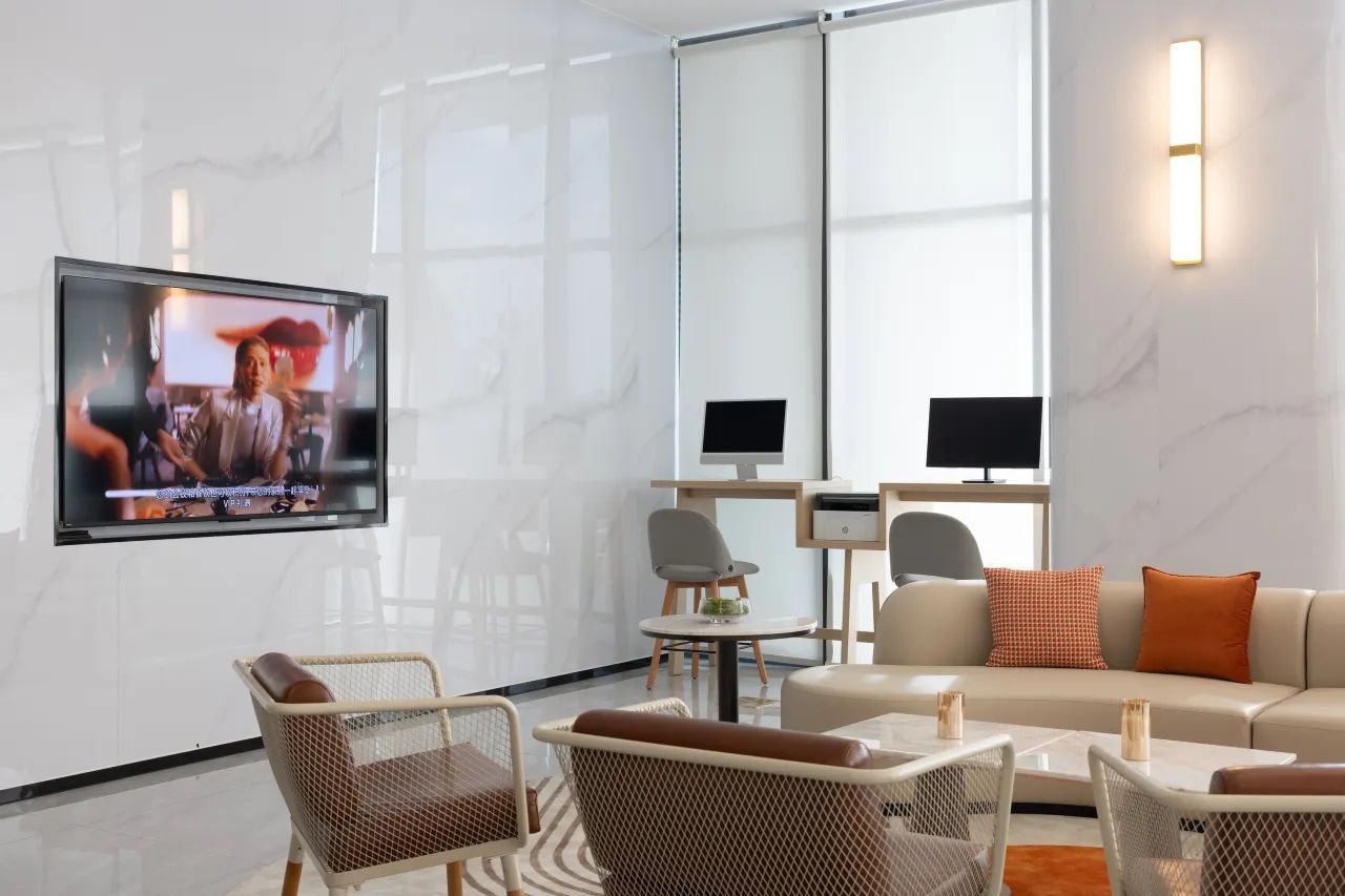 Een moderne hotellobby met een flatscreen televisie aan de muur, een bank, rieten stoelen, een klein tafeltje en een bureau met een computer.