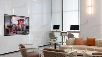 Een moderne hotellobby met een flatscreen televisie aan de muur, een bank, rieten stoelen, een klein tafeltje en een bureau met een computer.