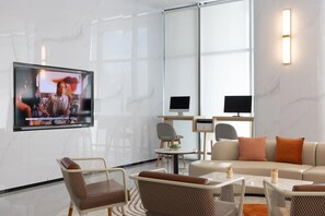 A modern hotel lobby with a flat-screen TV mounted on the wall, a sofa, wicker chairs, a small table, and a desk with a computer.