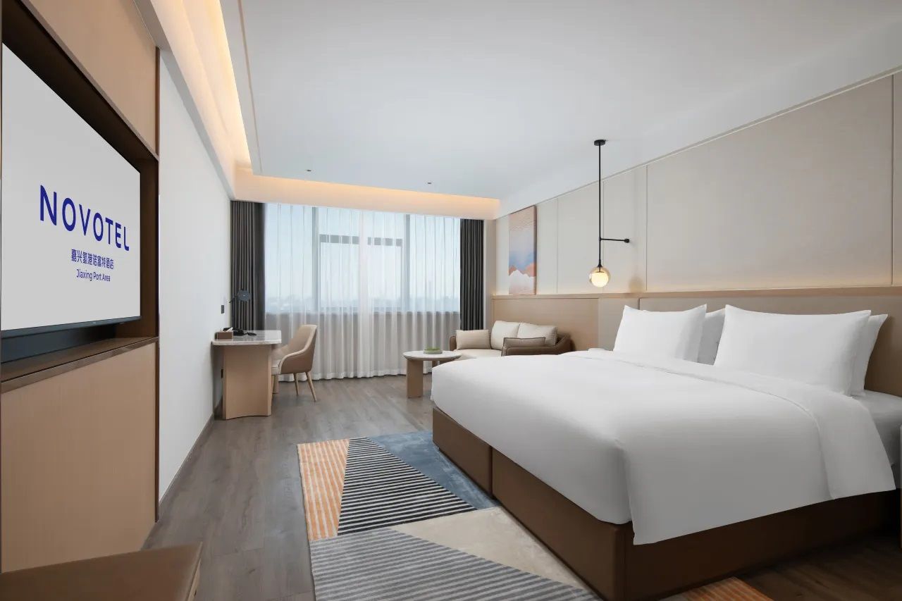 Una habitación de hotel moderna con una cama grande, un escritorio, una silla y una pantalla de televisión que muestra el nombre del hotel.
