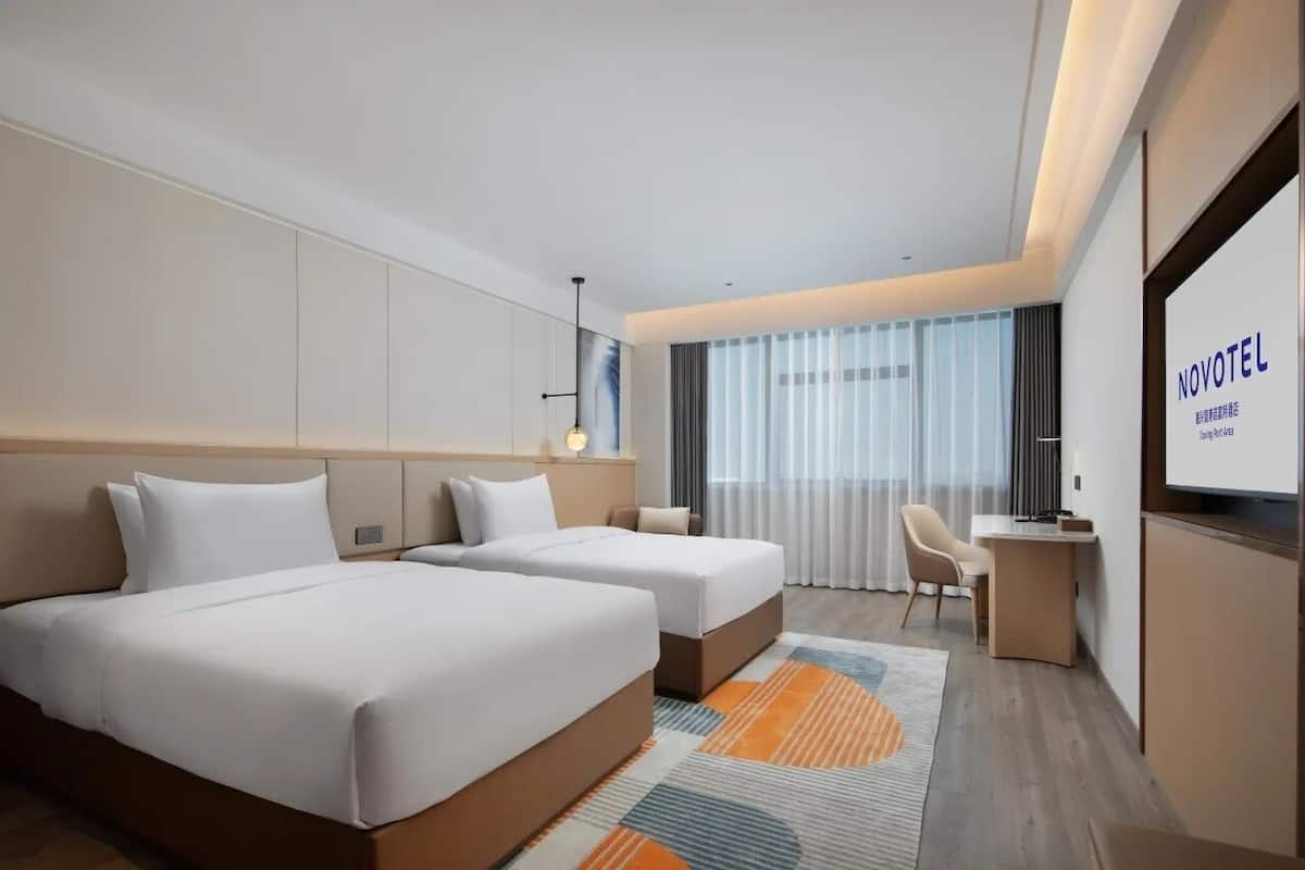A modern hotel room with two beds, a desk, and a large window with curtains.