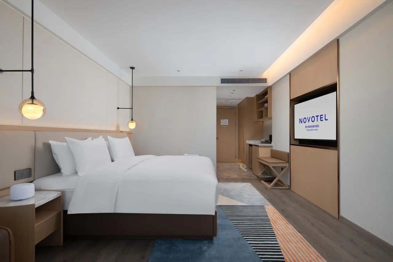 A modern hotel room with a large bed, bedside tables, a TV, and a view into the hallway.