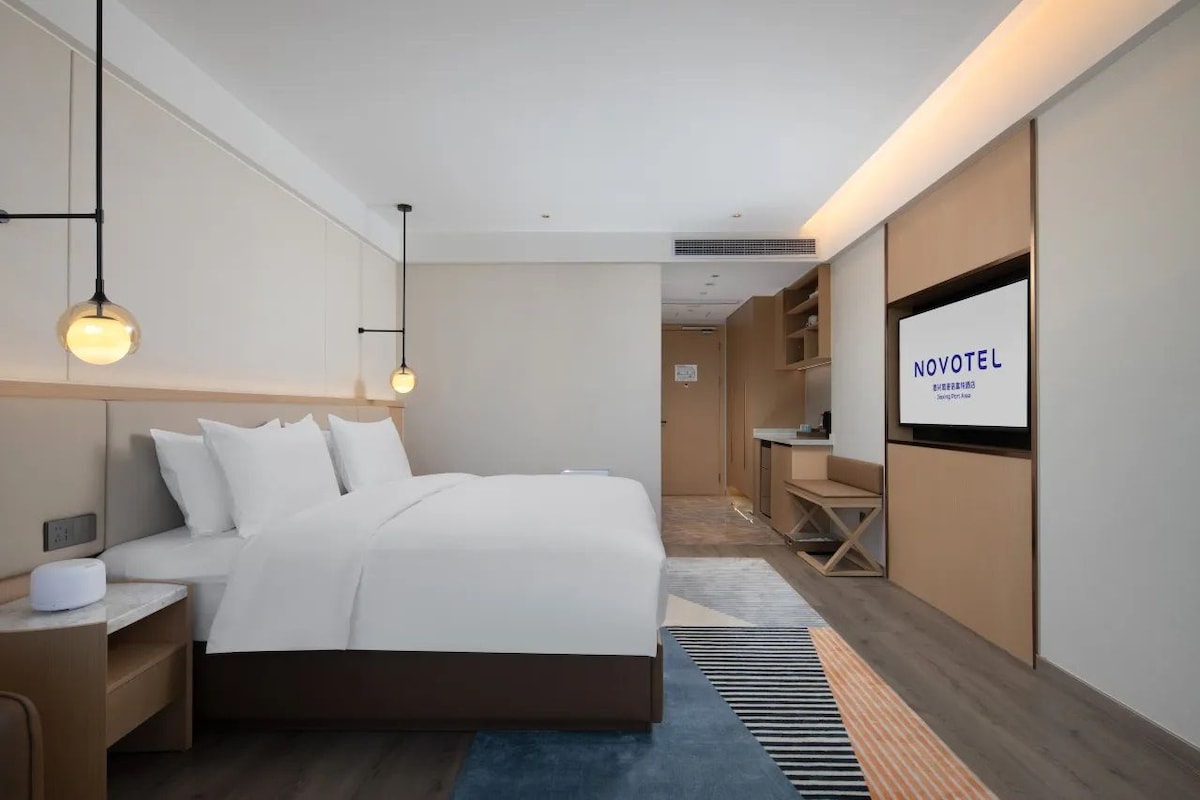 A modern hotel room with a large bed, bedside tables, a TV, and a view into the hallway.