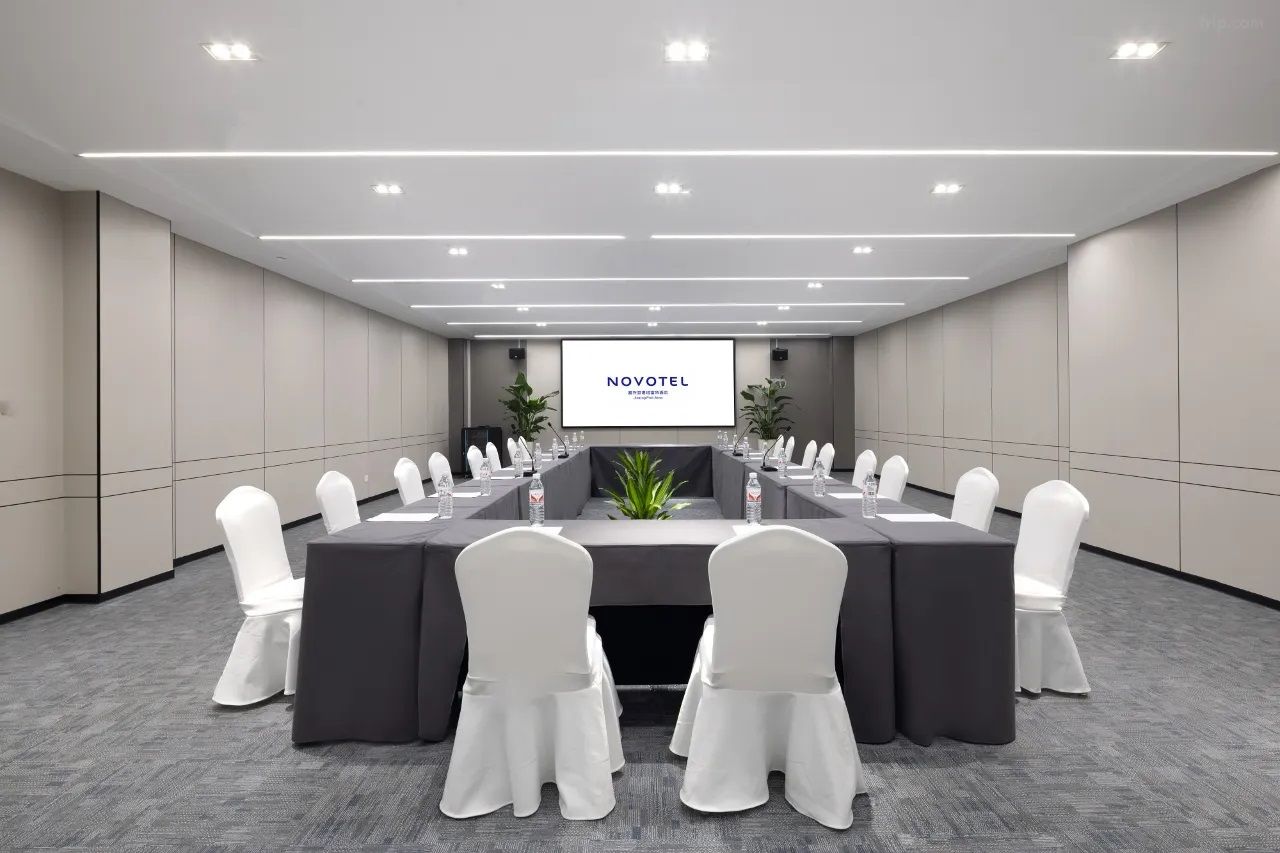 Un salón de conferencias con una larga mesa, sillas blancas y una pantalla que muestra "NOVOTEL".