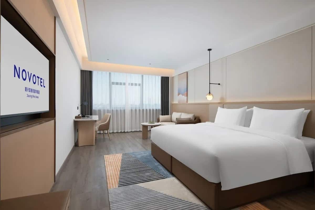A modern hotel room with a large bed, a desk, a chair, and a TV screen displaying the hotel name.