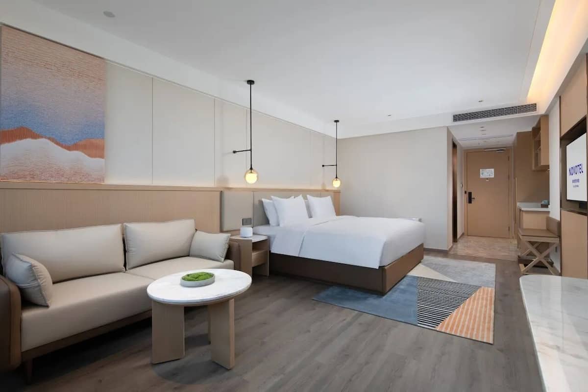 A modern hotel room with a bed, a sofa, a small table, and a painting on the wall.