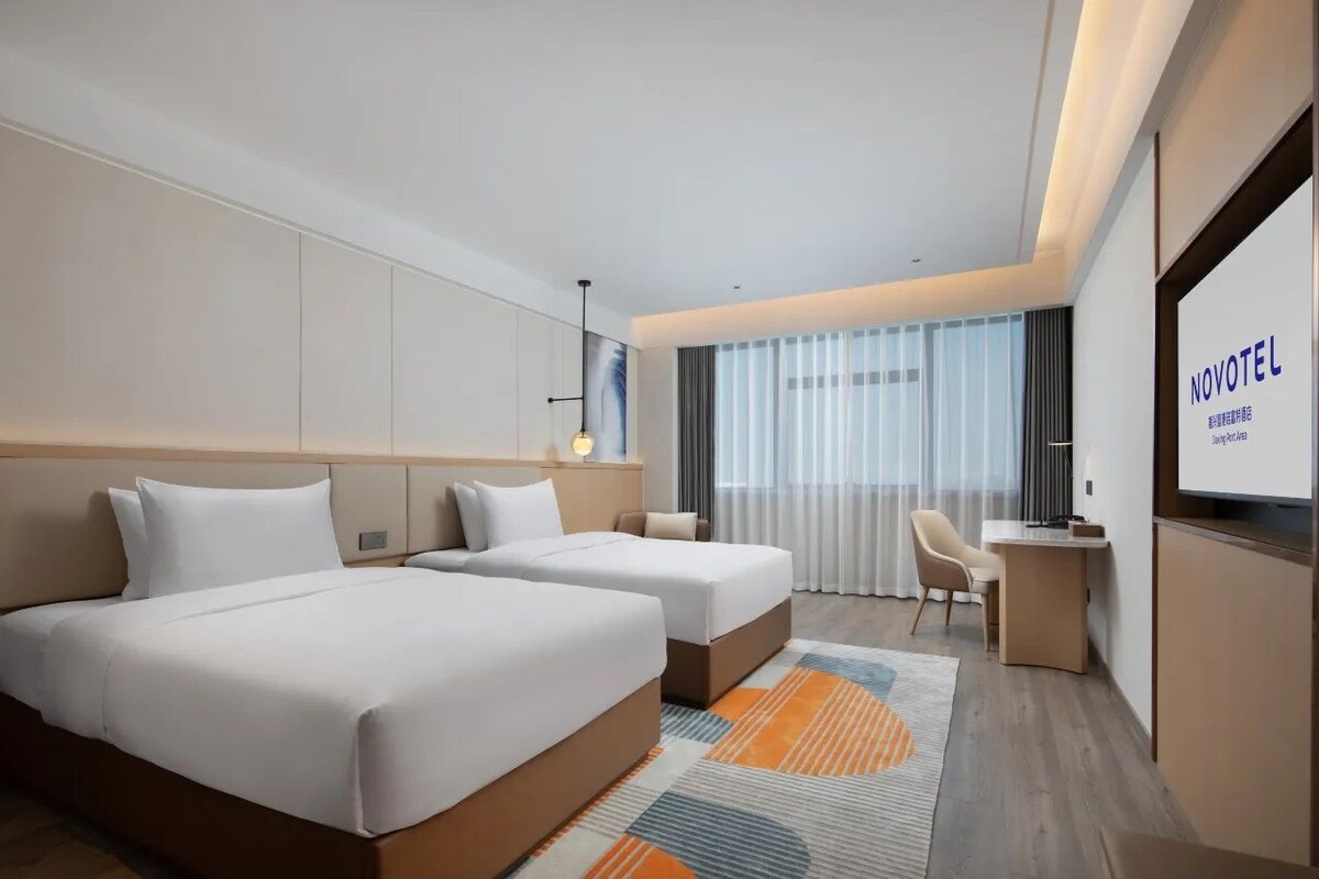 A modern hotel room with two beds, a desk, and a large window with curtains.