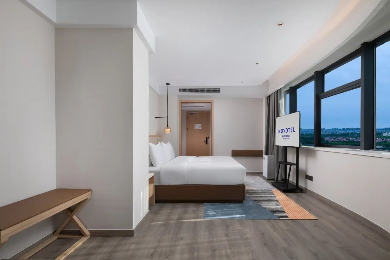 A modern hotel room with a large bed, a desk, and a view of the city through large windows.