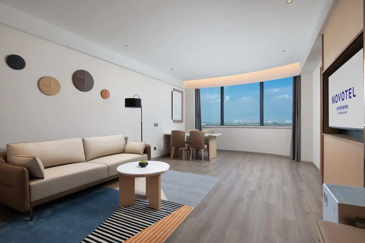 A modern hotel room with a sofa, a small round table, and a view of the city through large windows.