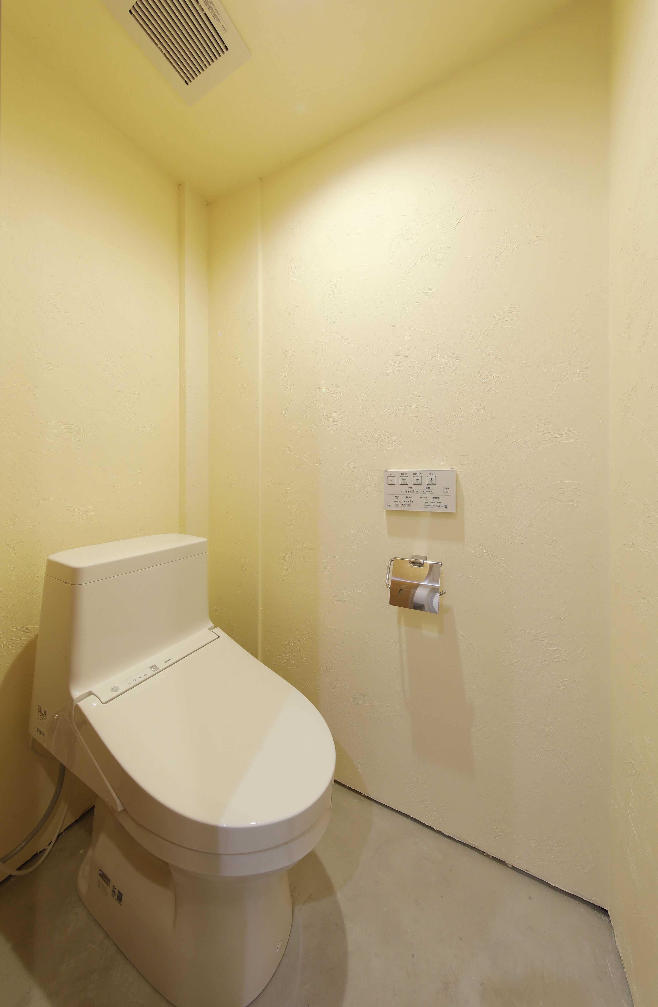 A small, plain bathroom with a white toilet, a wall-mounted flush handle, and a vent on the ceiling.