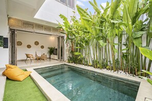 Un espace piscine intérieure moderne, doté d’un bassin aux eaux cristallines, entouré d’une végétation luxuriante et agrémenté d’un coin salon avec un coussin jaune.