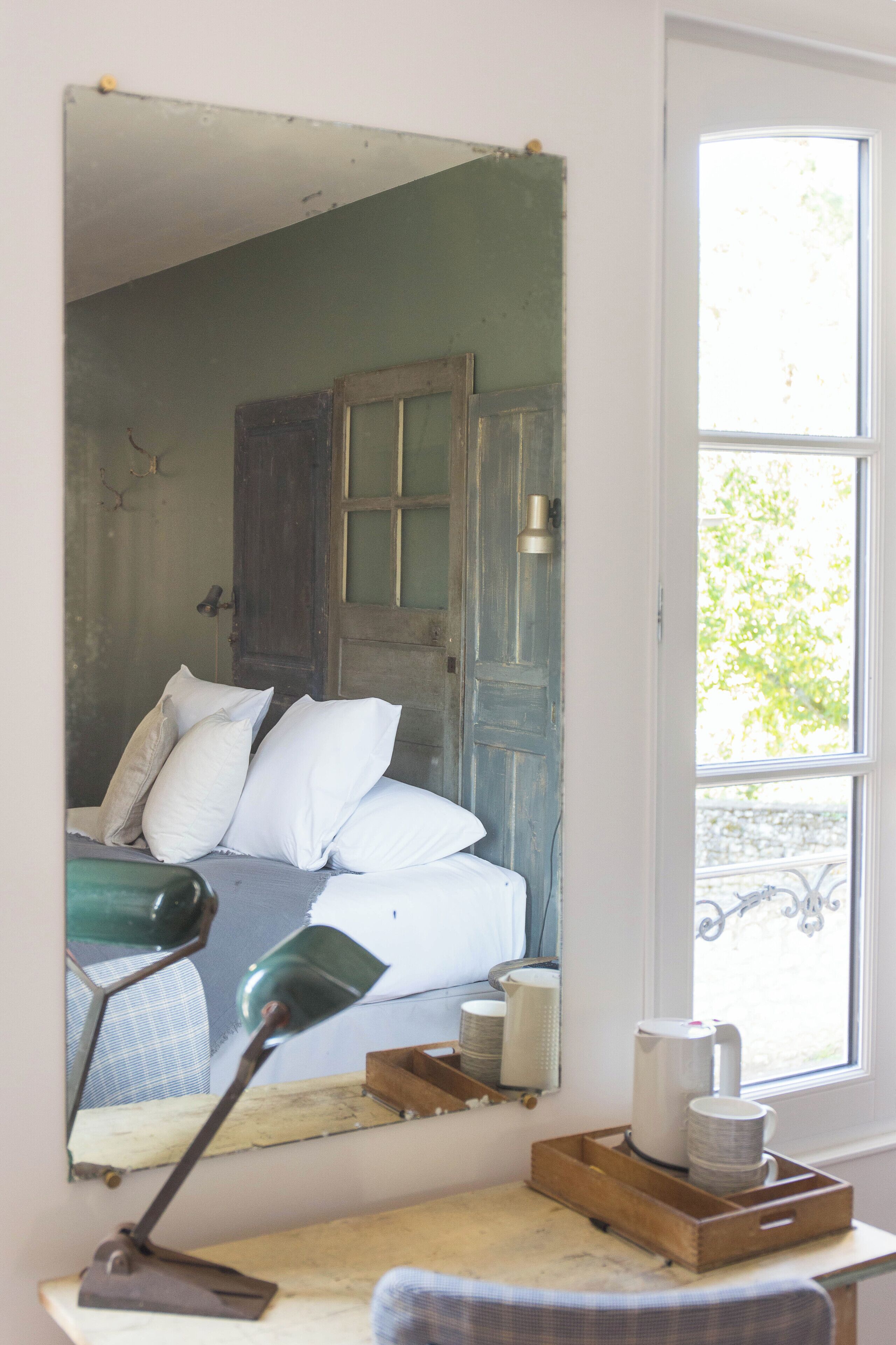A room with a bed, a desk with a lamp, a window, and a mirror reflecting the bed.