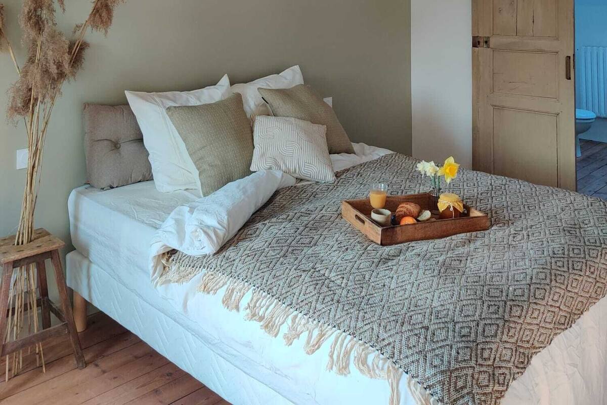 A bedroom with a bed, pillows, a bedside table, a wooden headboard, a hanging light fixture, and a view of the bathroom through an open door.