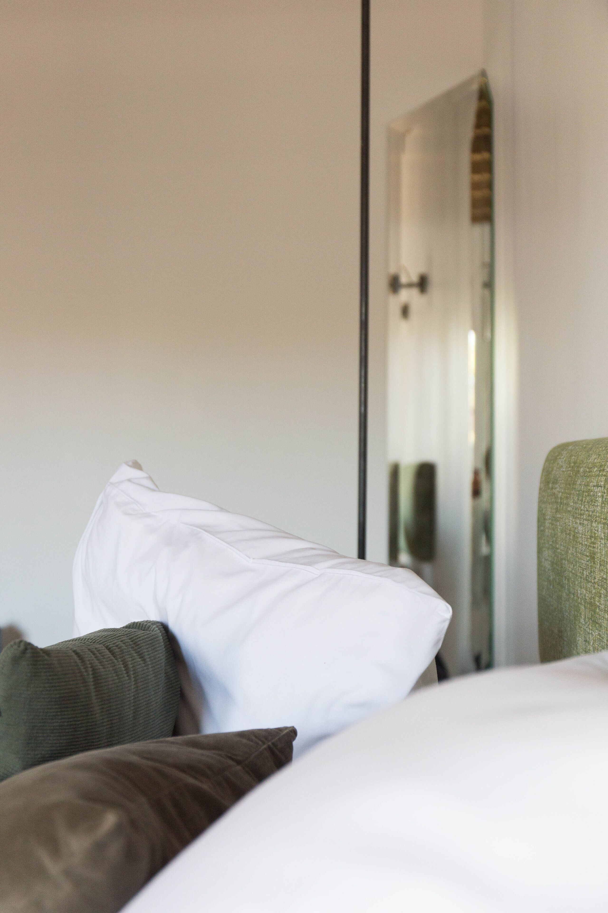 Habitación de hotel con cama, almohadas y un espejo que refleja una puerta y una lámpara.