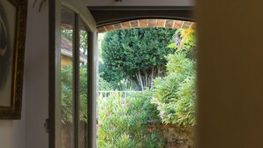 An open door leading to a patio with a table and chairs, a hanging bird sculpture, and a view of a garden with various plants. - maison seize (Perche-en-Noce)