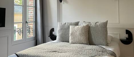 Un dormitorio con cama, almohadas, mesita de noche, lámpara de pared y ventana con cortinas.