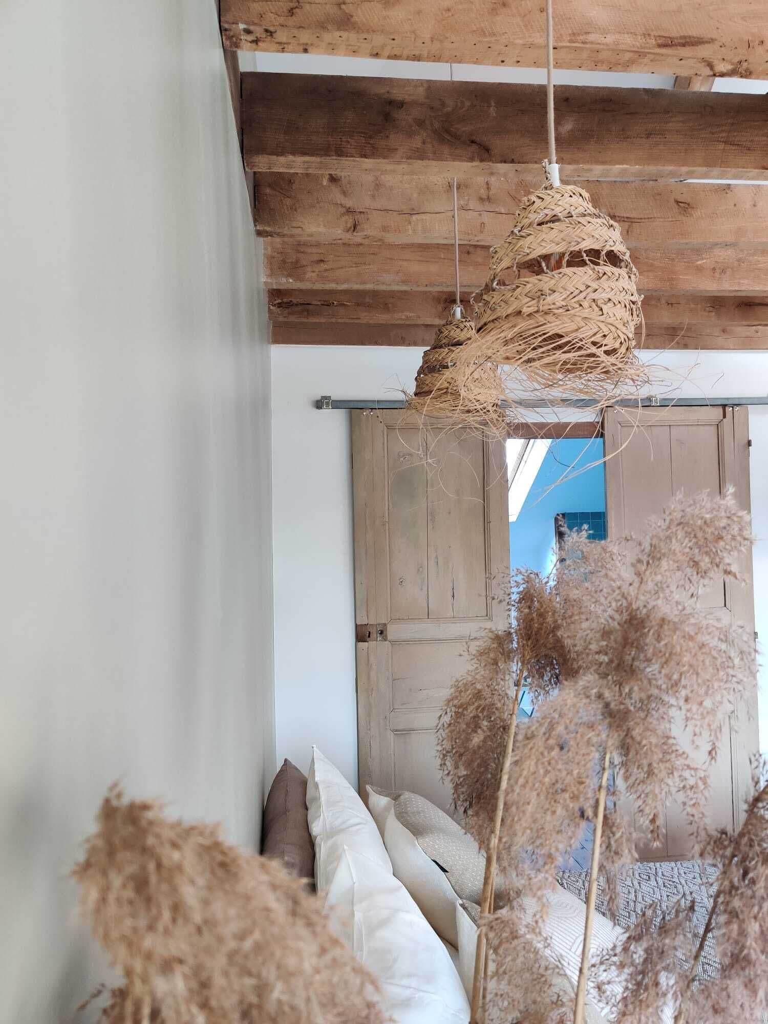 Ein rustikaler Raum mit einer gewebten Pendelleuchte, einer Holztür und einem Bett mit vielen Kissen.