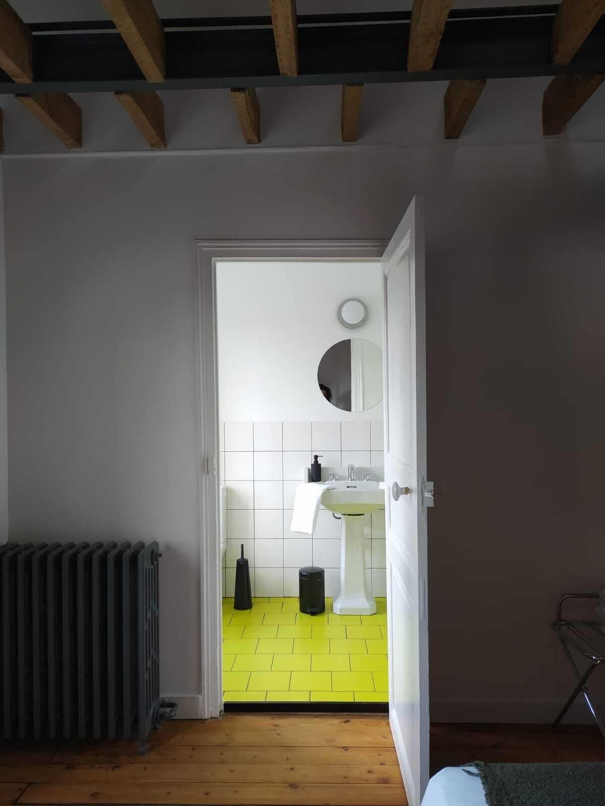 Quarto com chão de madeira, radiador e porta que dá para um banheiro com pia branca e espelho.
