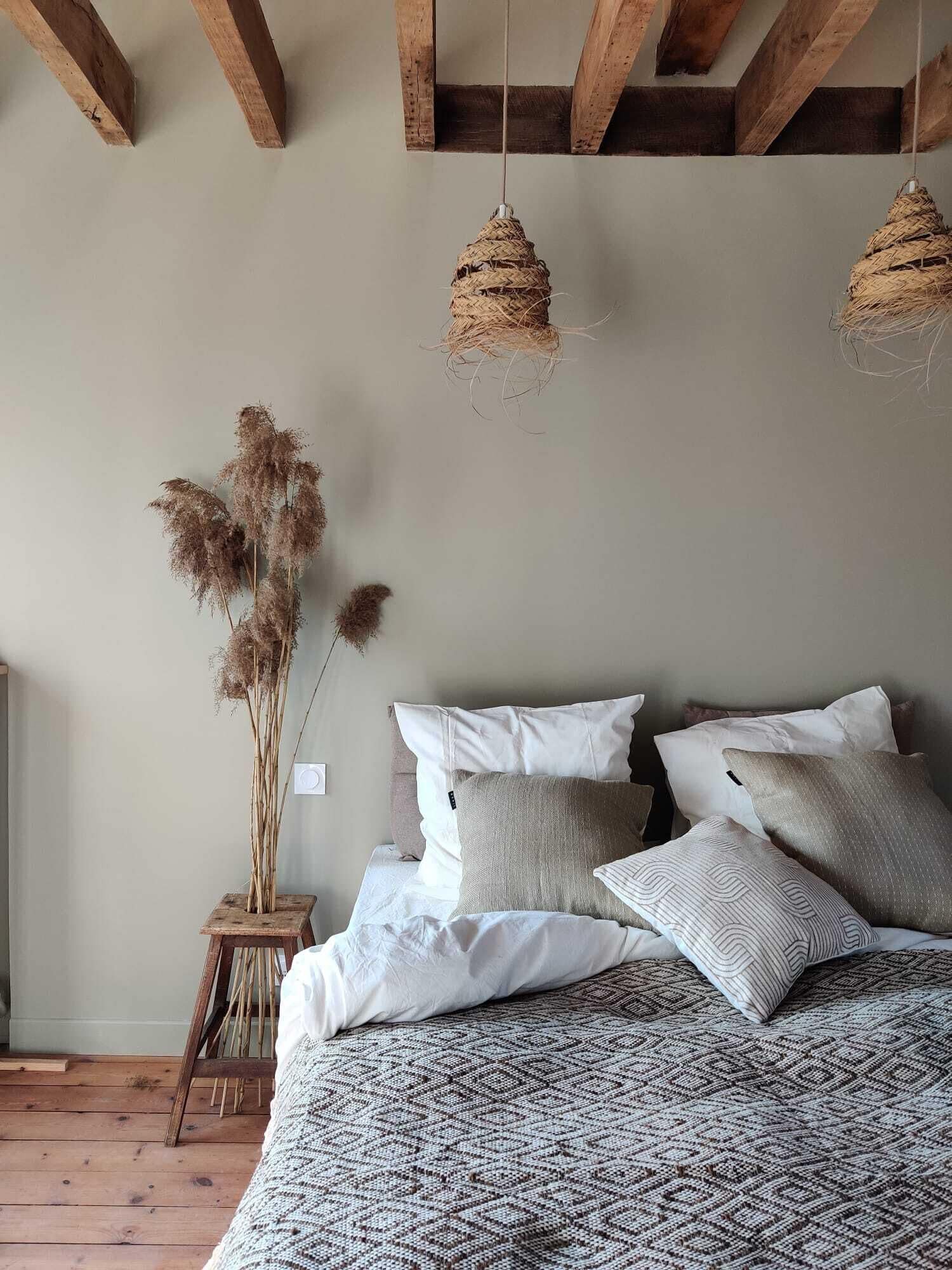 Un dormitorio con cama de madera, ropa de cama blanca y estampada, un pequeño taburete de madera con plantas secas, y lámparas de fibra.