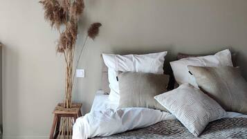 Un dormitorio con cama de madera, ropa de cama blanca y estampada, un pequeño taburete de madera con plantas secas, y lámparas de fibra.