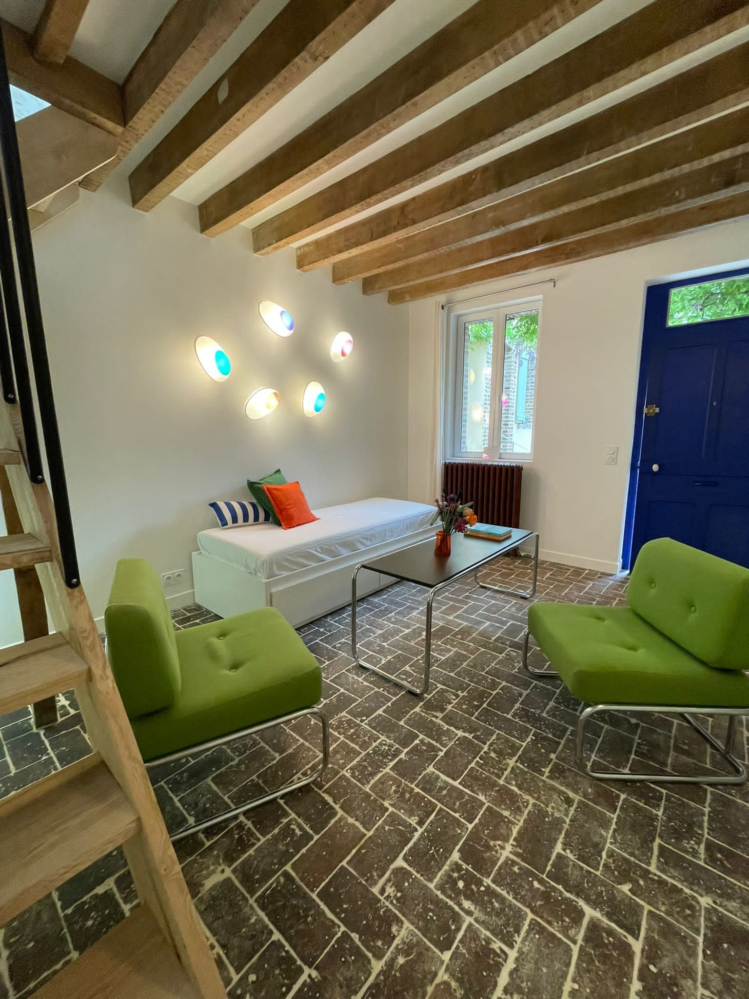 Un salon moderne avec un lit en mezzanine, des fauteuils verts, une table basse en verre et des poutres apparentes.