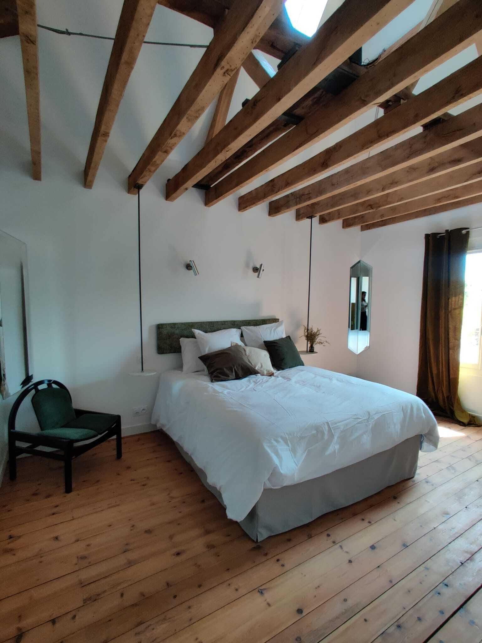 A bedroom with a wooden ceiling, a bed with white bedding, a chair, and a mirror on the wall.