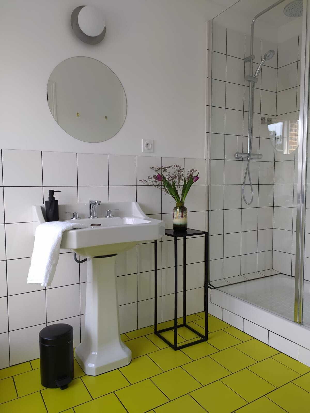 A bathroom with a white sink, a mirror, a vase with flowers, a black trash bin, and a shower area.