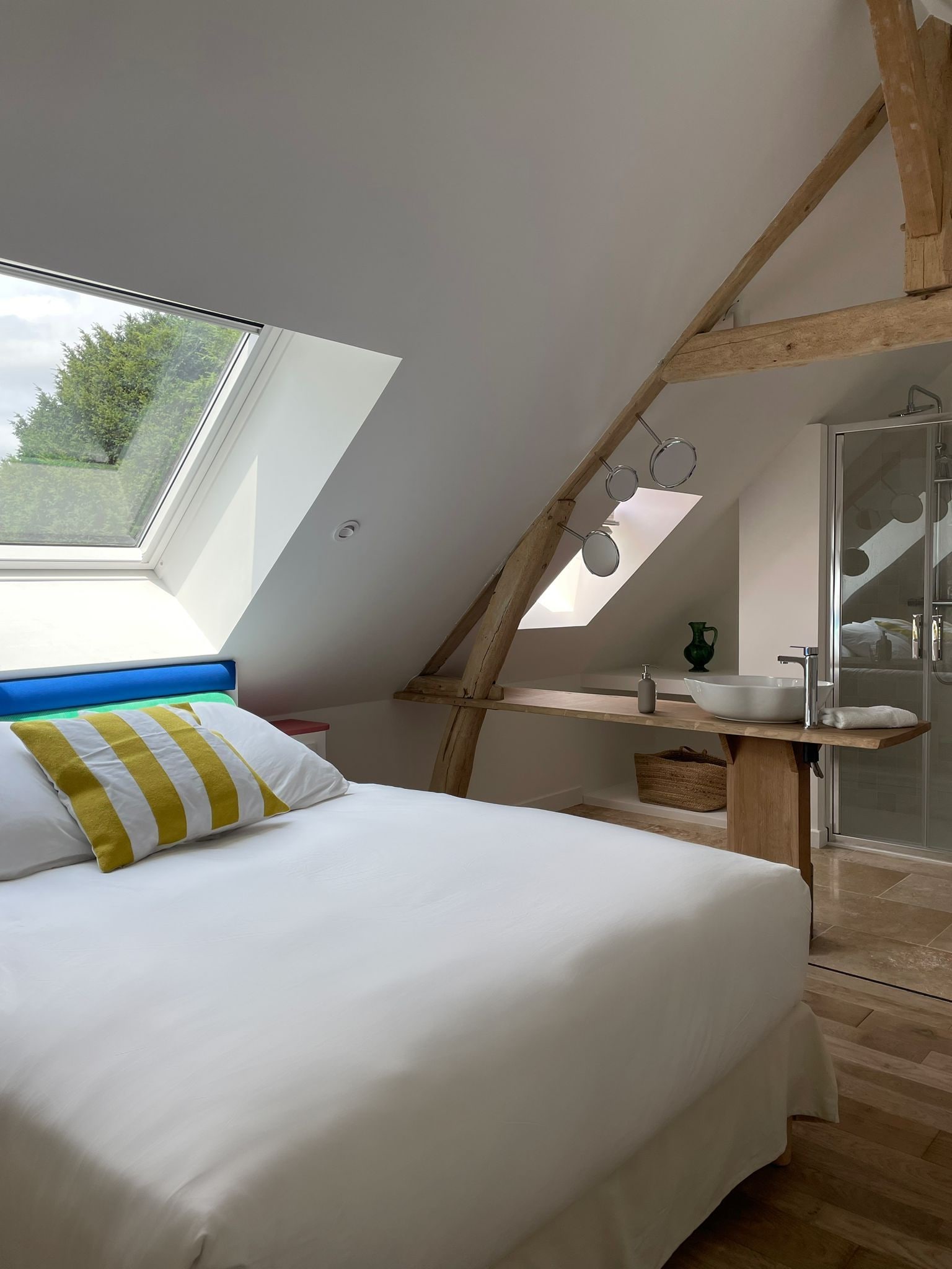 A bedroom with a skylight, a bed with striped pillows, a wooden shelf with toiletries, and a shower area.