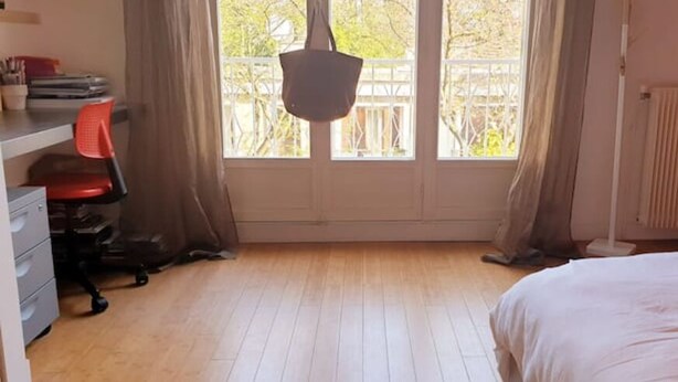 A bedroom with a bed, a desk with a chair, a radiator, and a window with curtains.