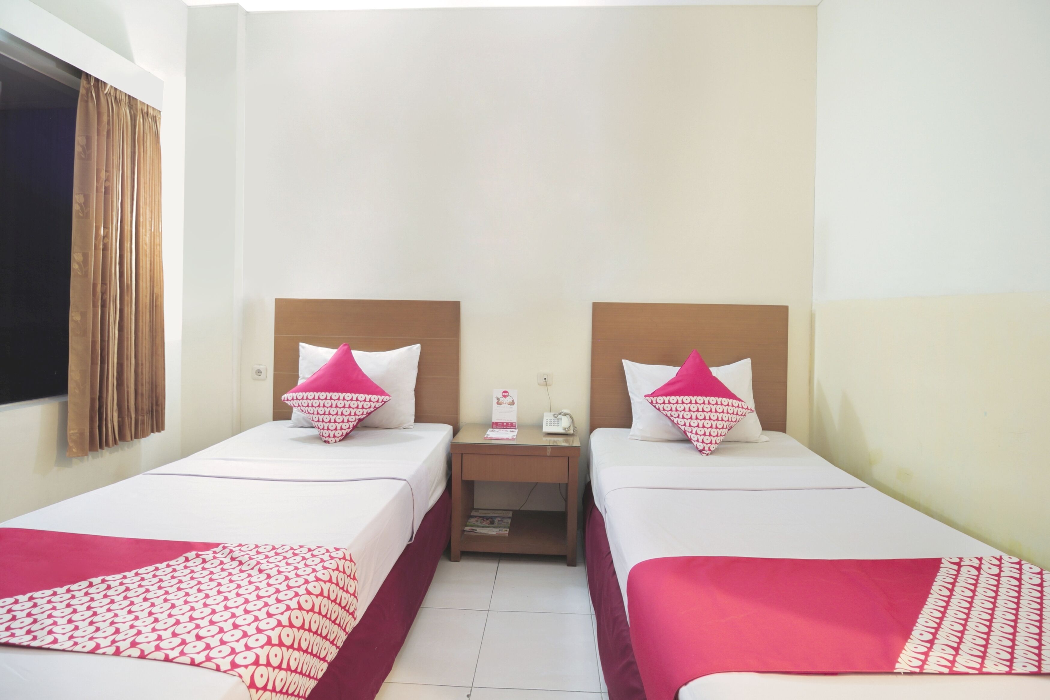 Une chambre d’hôtel avec deux lits simples, chacun doté d’une tête de lit en bois, une table de chevet entre les deux, et une fenêtre avec des rideaux.