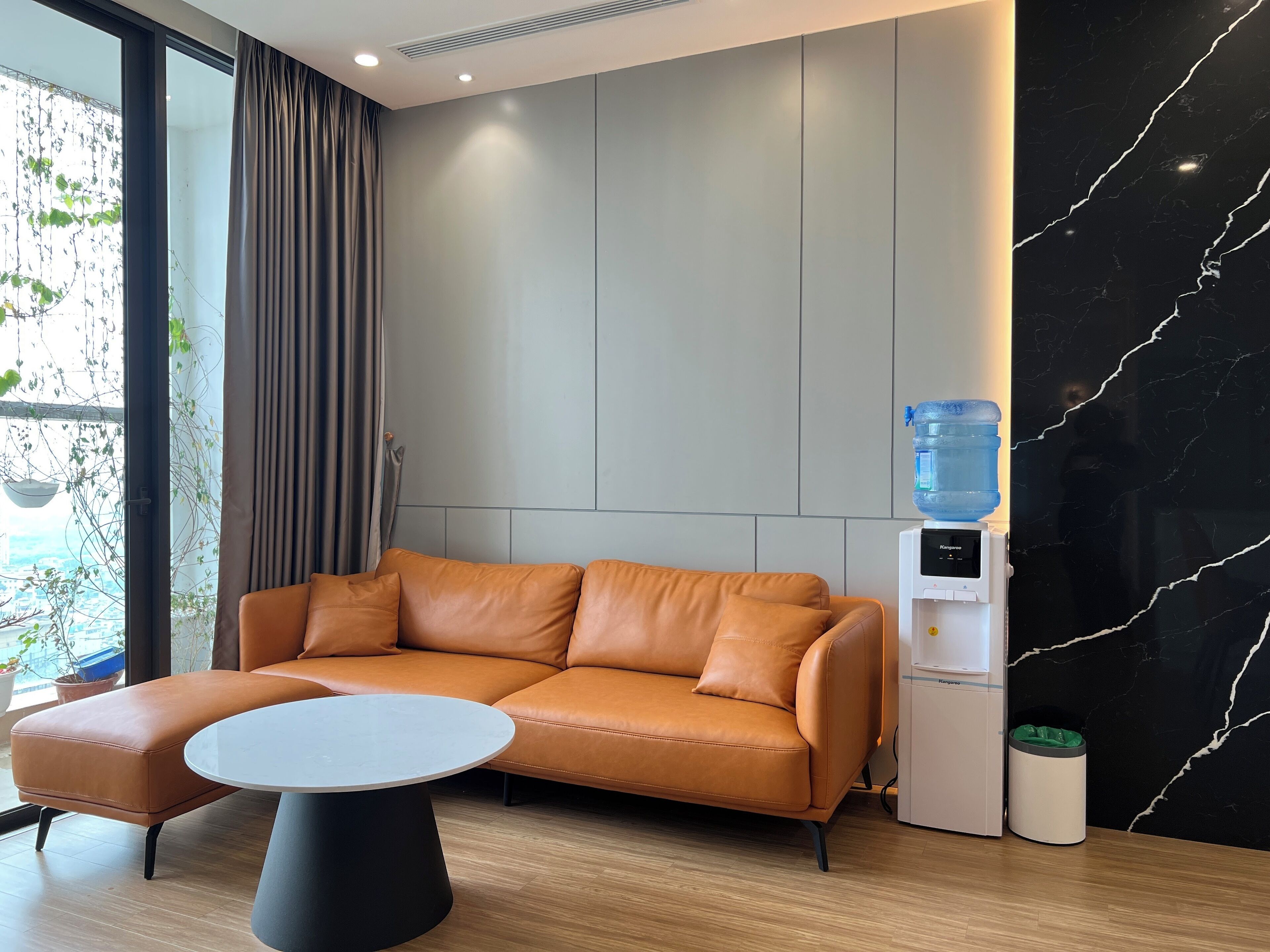 A modern room with a leather sofa, a round table, a water dispenser, and a view of the city through large windows.