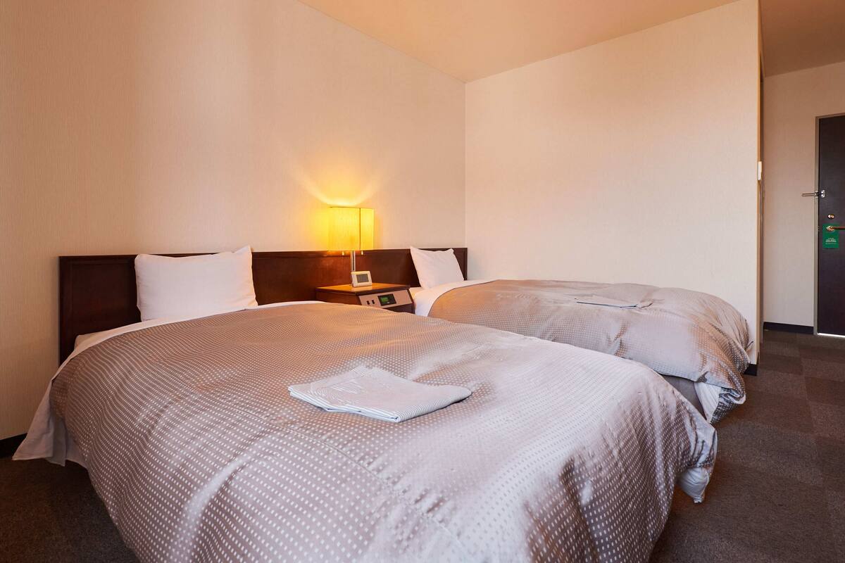 A hotel room with two beds, a nightstand with a lamp, and a door with a green sign.