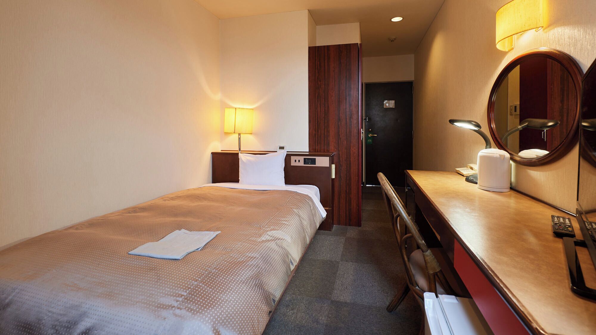 A hotel room with a bed, bedside table, lamp, mirror, and a desk with a telephone and a kettle.
