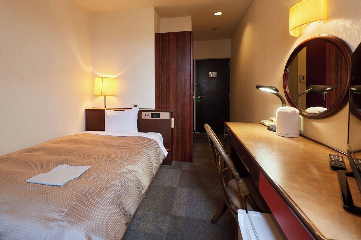 A hotel room with a bed, bedside table, lamp, mirror, and a desk with a telephone and a kettle.