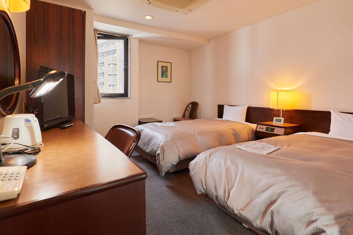 A hotel room with two beds, a desk with a telephone and lamp, a chair, and a window with a view of buildings.