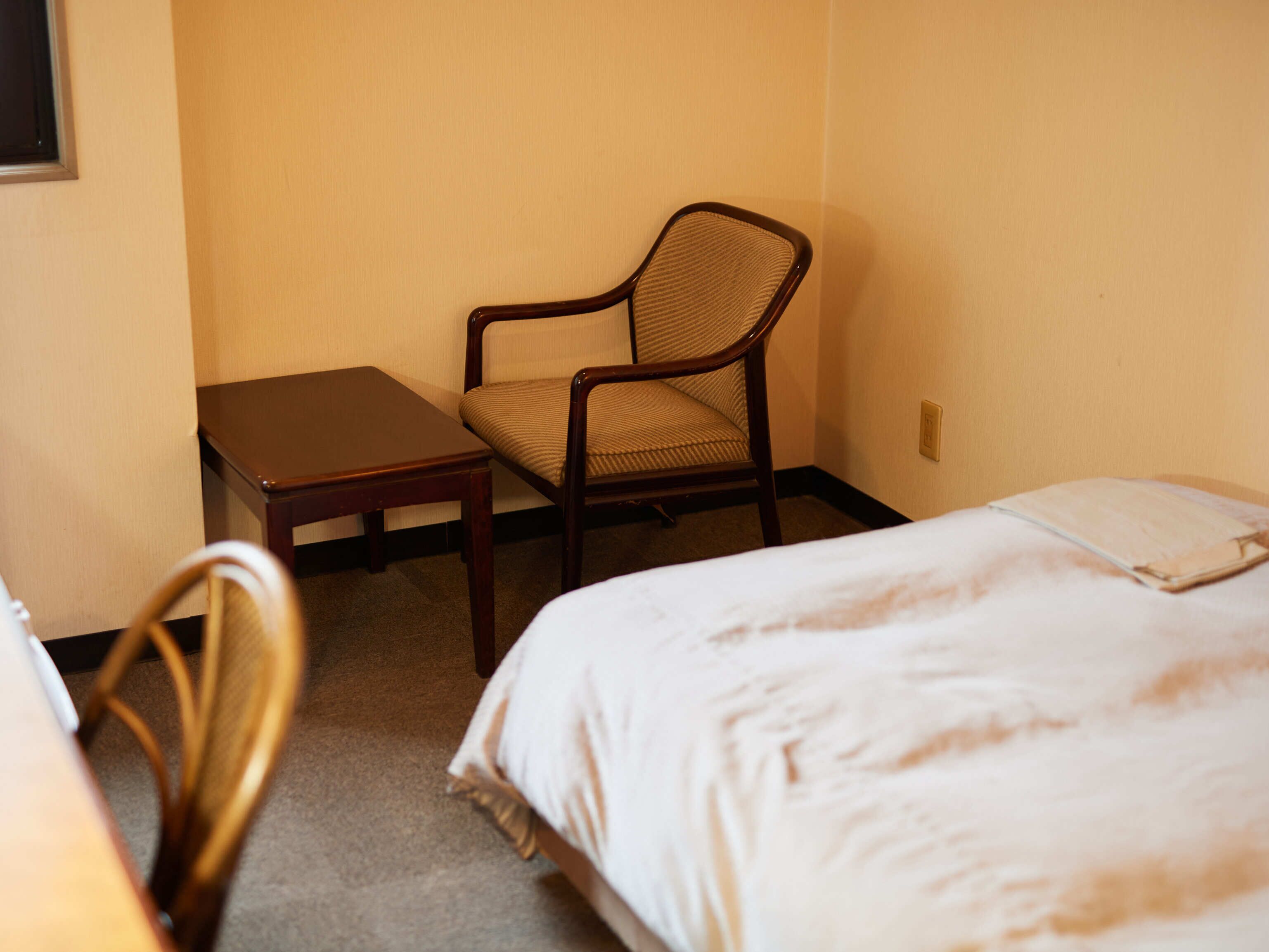 A hotel room with a single bed, a chair, a small table, and a wall socket.