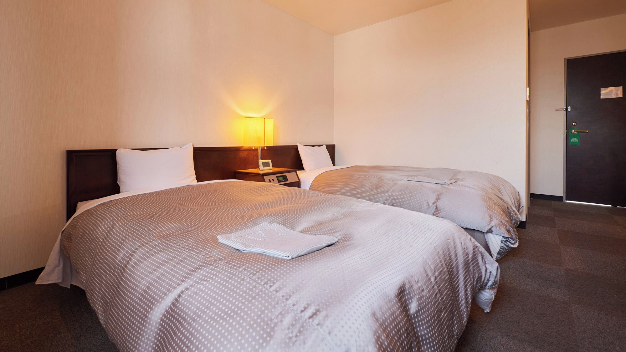 A hotel room with two beds, a nightstand, a lamp, and a door.
