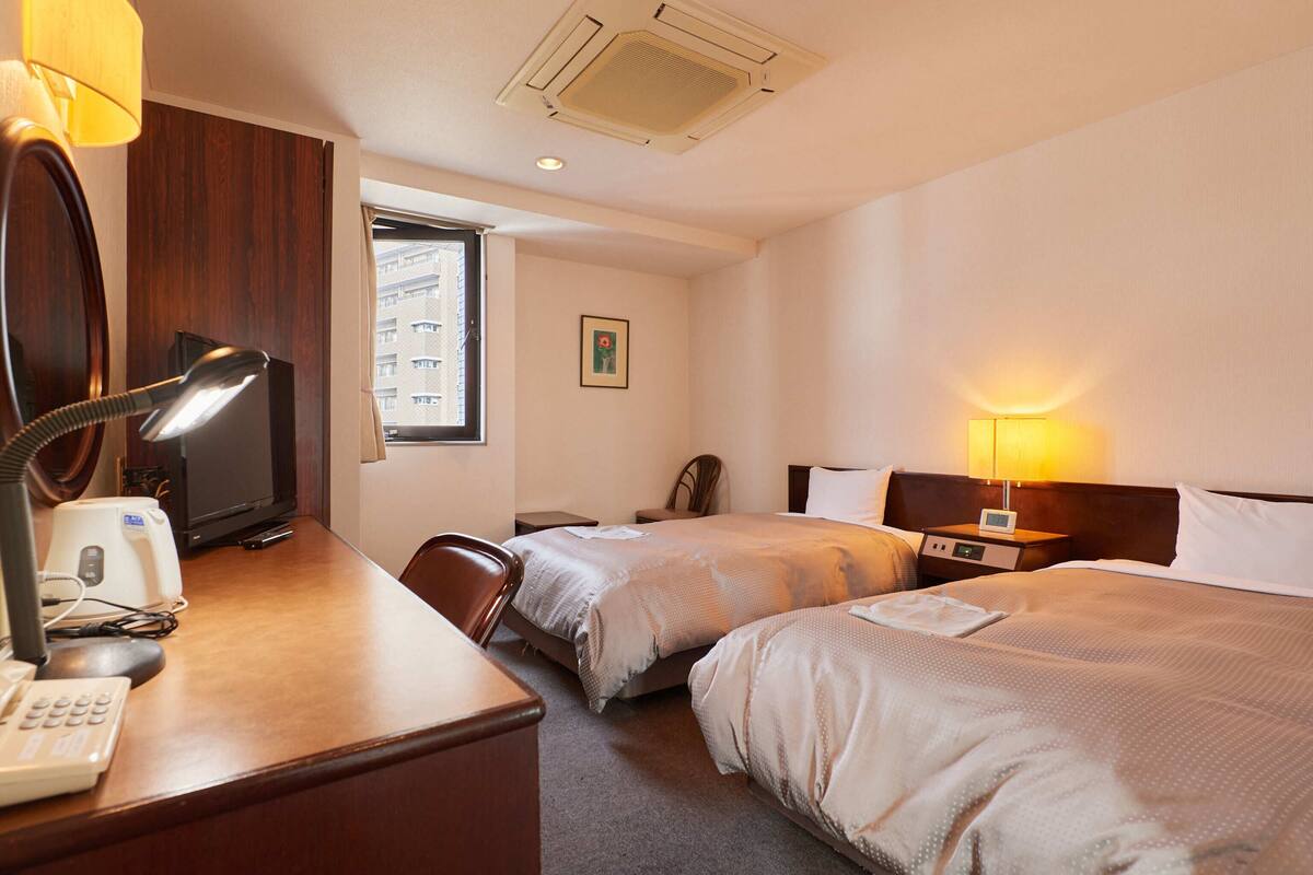 A hotel room with two beds, a desk with a lamp, a telephone, and a window with a view of buildings.