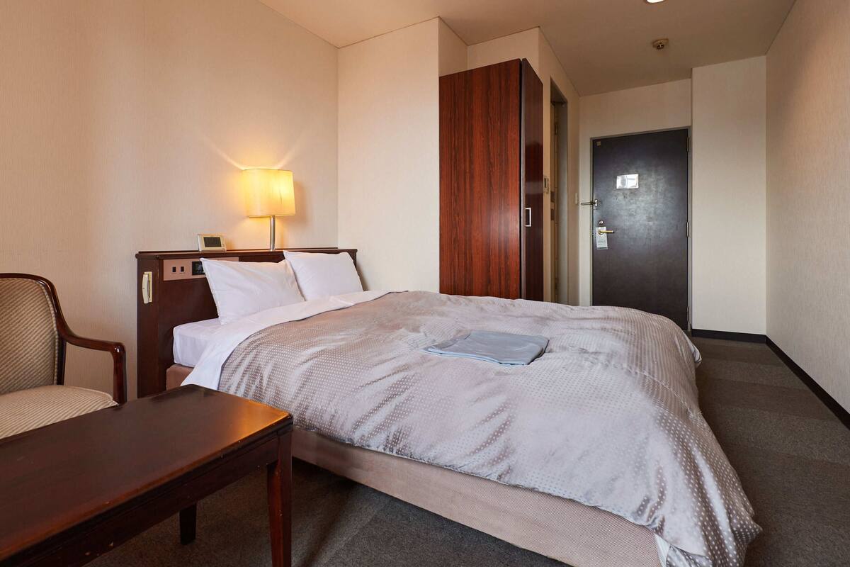 A hotel room with a bed, a desk, a chair, a lamp, and a door.