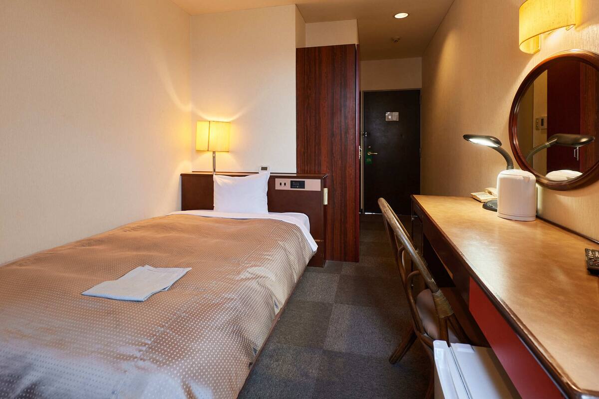 A hotel room with a bed, a desk, a chair, a desk lamp, a telephone, and a mirror.
