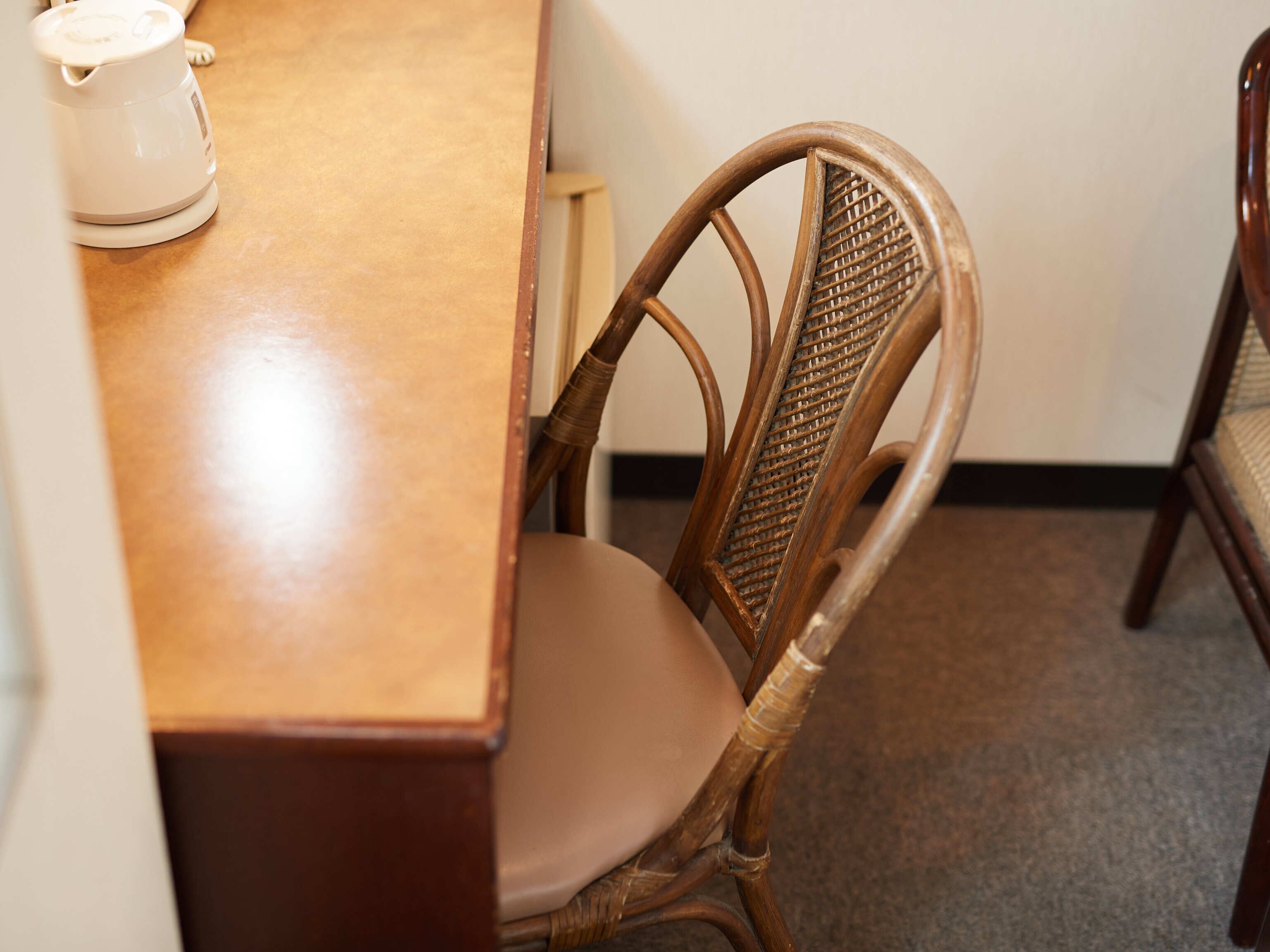 Una mesa de madera con un objeto blanco encima, una silla de mimbre con un cojín color crema, y un suelo alfombrado.