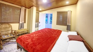 Een slaapkamer met een houten plafond, een bed met een rode deken, een rieten stoel en uitzicht buiten door een deur.