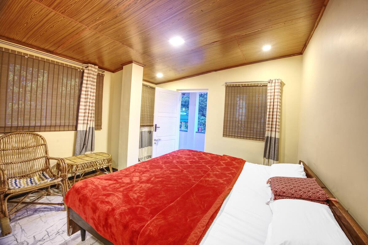 A bedroom with a wooden ceiling, a bed with a red blanket, a wicker chair, and a view of the outside through a door.