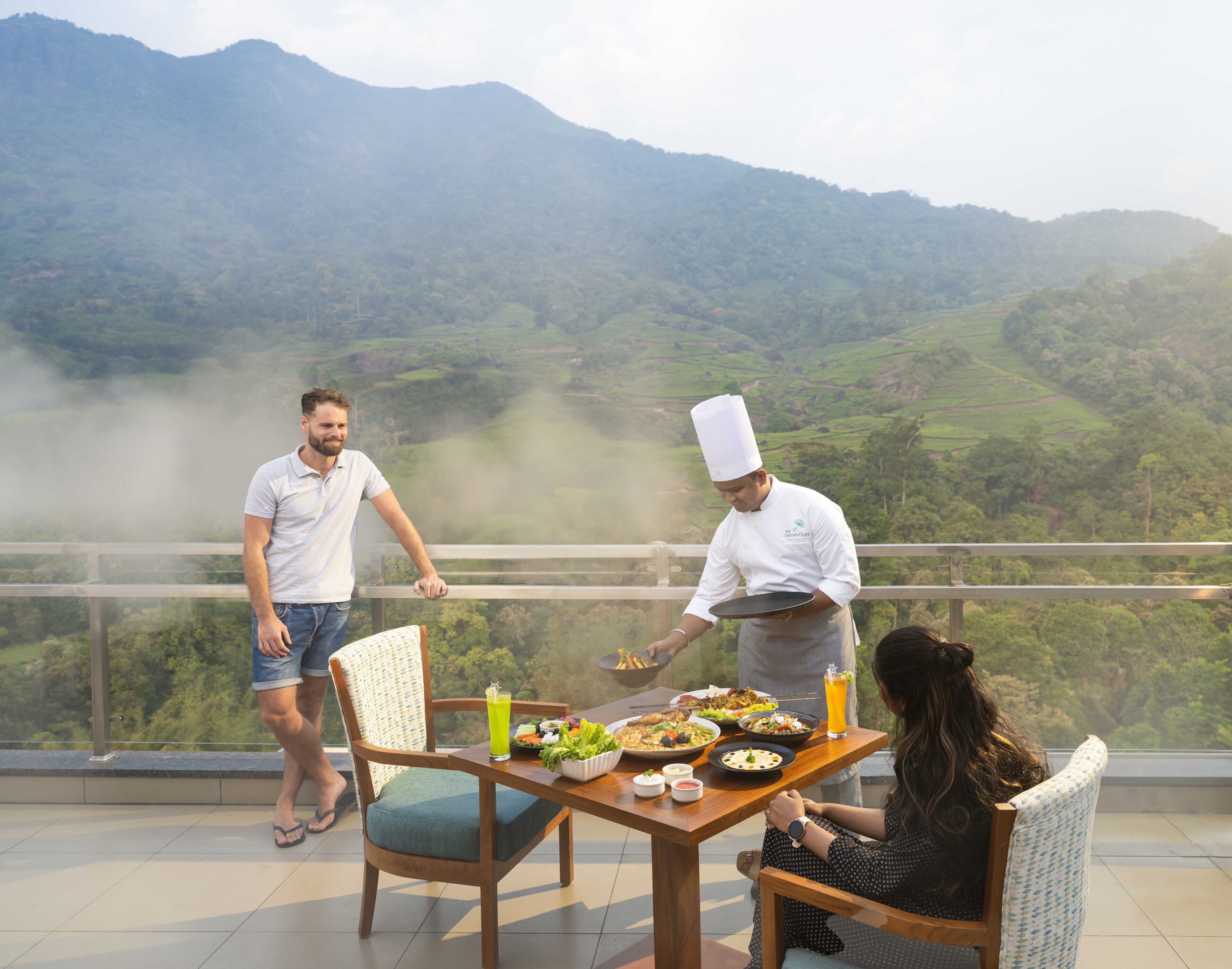 Une table à manger dressée avec différents plats, un chef qui sert les mets, et une vue panoramique sur les montagnes en arrière-plan.