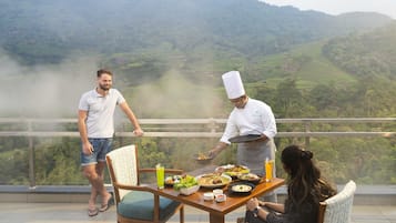 Une table à manger dressée avec différents plats, un chef qui sert les mets, et une vue panoramique sur les montagnes en arrière-plan.