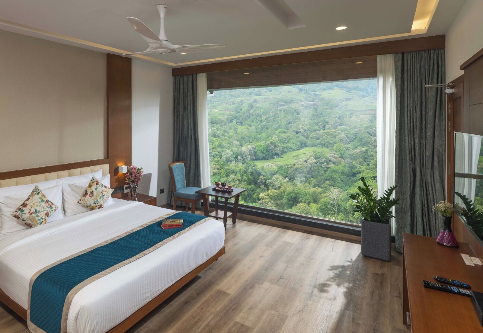 Habitación de hotel con una cama grande, un escritorio, una silla, un ventilador de techo y vistas a un paisaje boscoso.